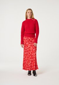 Rotes geripptes Sweatshirt mit hochgeschlossenem Kragen, kombiniert mit einem langen, gemusterten Rock in Rottönen und Pink, akzentuiert mit schwarzen Blumen. Schwarze Stiefeletten mit offenen Zehen.