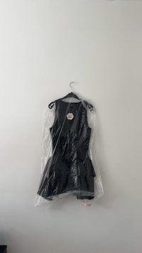 Schwarzes ärmelloses Kleid in einem transparenten Plastikkleidbeutel, an einem schwarzen Kleiderbügel aufgehängt. Auffällige Etiketten auf dem Kleid sichtbar. Glatte Textur.
