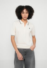 Polo de manga corta en color crema con cuello de cremallera, con detalles de rayas negras y un pequeño logo bordado en el pecho.