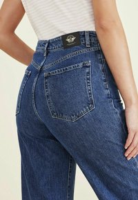 Vaqueros de denim azul de cintura alta con un corte relajado, que cuentan con dos bolsillos traseros y una etiqueta de la marca en la cinturilla. La textura es suave y duradera.