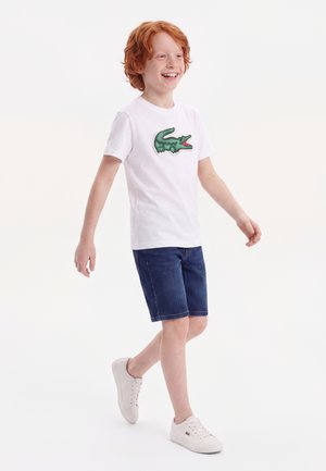 Garçon roux souriant marchant en t-shirt blanc avec un logo crocodile vert, short en jean et baskets blanches sur fond blanc.