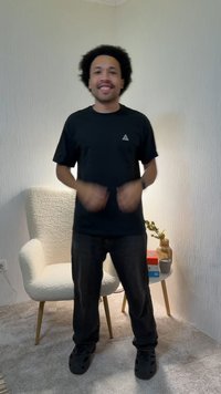 Mann med krøllete hår iført svarte klær smiler og gestikulerer, stående i et rom med teppe, en hvit lenestol og et lite bord med bøker og en vase.