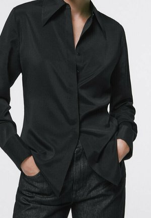 Personne portant une chemise noire à manches longues boutonnée avec col, rentrée dans un pantalon noir taille haute, mains dans les poches.