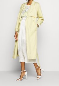Lys gul trenchcoat med dobbeltrække front, parret med en blomsterprint-top og en hvid plisseret nederdel. Accessoireret med hvide højhælede sko.
