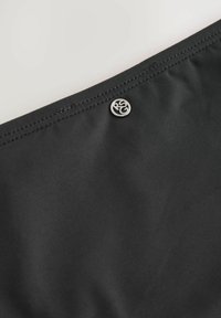 Črn material z gladko teksturo, s srebrnim gumbom logotipa na zgornjem robu in kontrastnim šivanjem ob šivu.