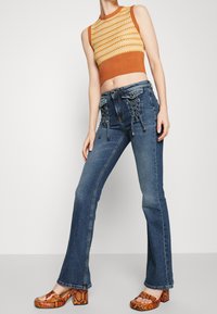 Hnědý a žlutý pletený crop top v kombinaci s tmavě modrými rozšířenými džíny s šněrovacími detaily a oranžovými platformovými lodičkami se vzorem hada.