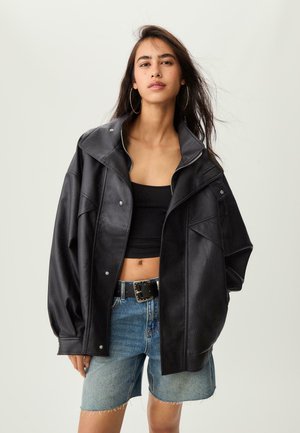 Ung kvinde iført en oversized sort læderjakke, sort crop top, hoops øreringe, stålbesat sort bælte og denim shorts med flosset kant.