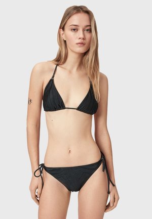 Ung kvinde iført en sort trekantsbikini med tynde stropper og sidebind, stående mod en enkel lys baggrund.