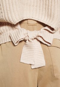 Maglione lavorato a maglia di colore crema con una texture a costine e un dettaglio con fiocco legato in vita, abbinato a pantaloni beige con tasca sul retro.