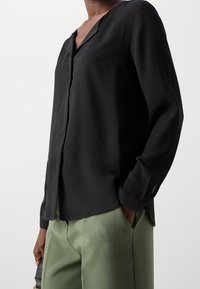Blusa nera a maniche lunghe con scollatura a V e vestibilità rilassata, realizzata in tessuto leggero; abbinata a pantaloni verde chiaro.