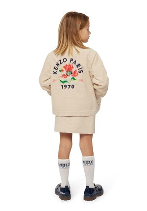 Beige corduroy jasje met bloemborduurwerk, tekst "KENZO PARIS 1970", gedragen over een beige rok, gecombineerd met witte sokken en donkere schoenen.