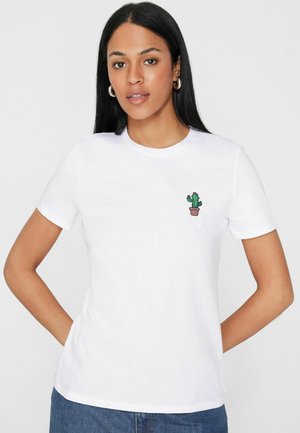 Vrouw met lang zwart haar, zilveren creolen oorbellen, een wit t-shirt met een kleine cactusborduursel en blauwe spijkerbroek, staand met handen achter zich.