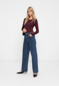 Donna bionda che indossa un top viola a maniche lunghe, jeans a vita alta e ampia blu, tacchi neri e una piccola borsa a tracolla nera.
