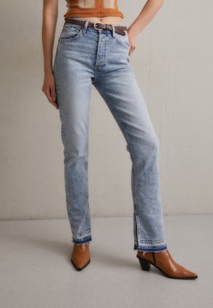 Frau trägt hellblaue High-Waist-Jeans mit ausgefransten Säumen und braune Stiefeletten mit Absatz, steht mit einer Hand auf der Hüfte.