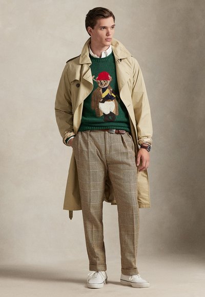 Μπεζ παλτό τύπου trench πάνω από πράσινο πουλόβερ με σχέδιο αρκούδας, γκρι καρό παντελόνι και λευκά αθλητικά παπούτσια. Ορατές λεπτομέρειες ζώνης και ρολογιού.