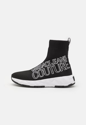 Baskets chaussettes montantes noires avec semelle blanche épaisse et grand texte blanc "Versace Jeans Couture" sur le côté.