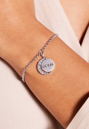 Zilveren armband met ketting aan de pols, versierd met een halve maan met kristallen en een ronde hanger waarop de merknaam "GUESS" gegraveerd is.
