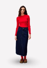 Rotes geripptes Langarmoberteil kombiniert mit einem maxirock aus dunklem Denim, der diagonale Nähte aufweist, getragen mit roten Socken und Clogs mit Leopardenmuster.