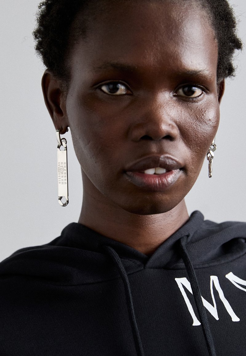 Svart hettegensere med hvit tekst. Sølvøredobb formet som en sikkerhetsnål, med gravert detaljer, båret på mørk hud.