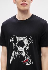 T-shirt de algodão preto com uma estampa branca da cara de um cão e um rótulo vermelho "HUGO" na área do colarinho. Mangas curtas, corte clássico.