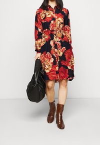 Abito a stampa floreale con fiori rossi e gialli su sfondo blu navy, con abbottonatura frontale, vita cinchata, maniche lunghe, abbinato a stivali alla caviglia marroni e una borsa nera.