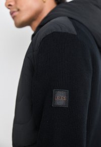 Pull en tricot noir avec un panneau d'épaule rembourré gris et une capuche ; présente une étiquette rectangulaire "BOSS" sur la manche gauche.