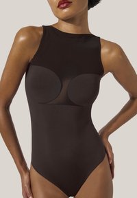 Kvinna som bär en ärmlös svart body med genomskinliga nätpaneler över bröstet, poserar med en hand på höften.