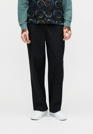 Asmuo dėvi laisvus juodus kelnes, baltus sportinius batelius ir žalią ilgomis rankovėmis marškinį su raštuotu juodu liemeniu, stovintis priešais paprastą foną.