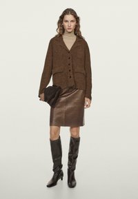 Donna che indossa un cardigan marrone peloso, un dolcevita beige, una gonna in pelle marrone testurizzata e stivali neri fino al ginocchio, mentre tiene una borsa piegata scura.