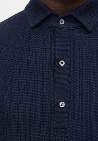 Gros plan sur un polo en tricot côtelé bleu foncé avec trois boutons blancs et un col plié sur une peau foncée.