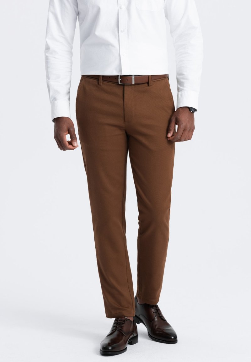 Braune Anzughose aus glattem Stoff, slim fit, mit Gürtelschlaufen und eingeknöpftem Vorderteil. Getragen mit einem weißen Hemd und dunkelbraunen Schuhen.