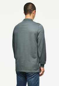 Muž s krátkými tmavými vlasy, oblečený v šedém polo tričku s dlouhým rukávem a modrých džínách, stojící zády k prostému pozadí.