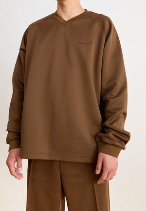 Mand iført brun Carhartt V-hals sweatshirt og matchende brune bukser mod en ensfarvet baggrund.