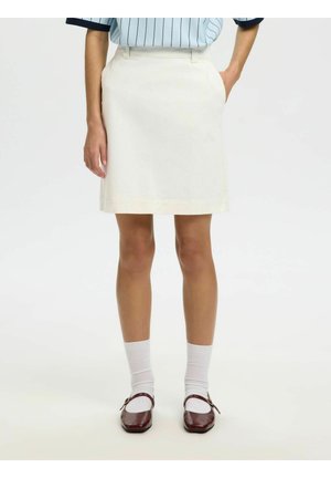 Femme portant une jupe blanche jusqu'aux genoux, une chemise rayée bleu clair, des chaussettes blanches et des chaussures Mary Jane bordeaux, debout les mains dans les poches.