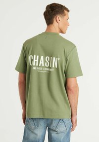 Žalias medvilninis marškinėliai trumpomis rankovėmis, kuriuose ant nugaros yra didelis baltas grafiškas tekstas. Dėvimi su mėlynomis džinsinėmis kelnėmis.