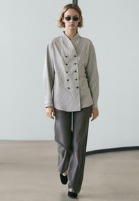 Gestreiftes Hemd mit Knöpfen und Stehkragen, kombiniert mit grauen, weit geschnittenen Hosen und schwarzen Ballerinas, getragen mit einer dunklen Sonnenbrille.