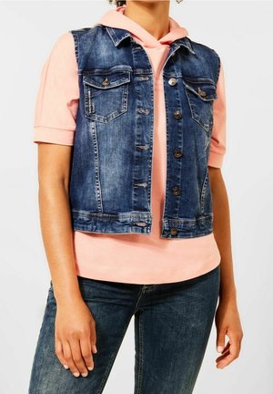 Personne portant un gilet sans manches en jean bleu sur un sweat à capuche rose clair et un jean bleu foncé, debout les bras détendus le long du corps.