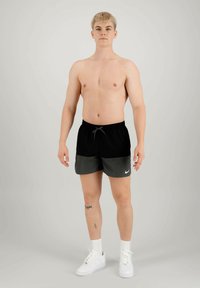 Shorts da bagno neri e grigi con vita regolabile, caratterizzati da un logo Nike bianco. Indossati con scarpe da ginnastica bianche e calzini bianchi alla caviglia.