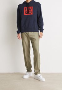 Persona vistiendo una sudadera azul marino con un logo cuadrado rojo, pantalones beige y zapatillas blancas, de pie sobre un suelo neutro frente a una pared blanca.