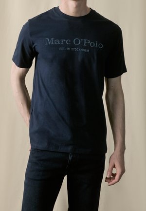 Homme portant un T-shirt noir Marc O'Polo à manches courtes et un jean noir devant un fond beige uni.