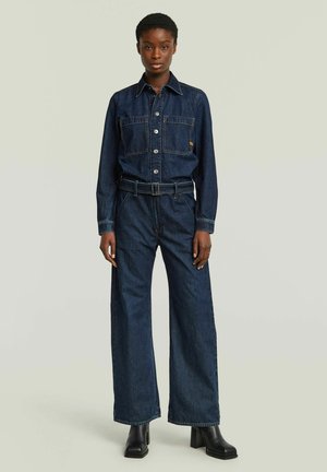 Junge Frau, die einen dunkelblauen Denim-Overall mit langen Ärmeln und weitem Hosenbein trägt, kombiniert mit schwarzen Stiefeln mit Absatz.