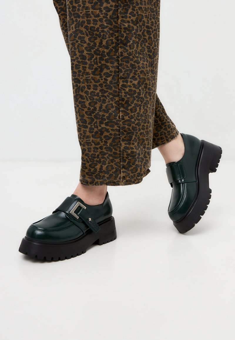 Chaussures compensées en cuir vert foncé avec semelles épaisses noires et boucle argentée, portées avec un pantalon court marron à imprimé léopard.