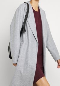 Donna che cammina indossando un lungo cappotto grigio chiaro sopra un vestito bordeaux, con una borsa nera a tracolla con cuciture bianche.