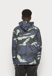 Giacca da pioggia camouflage nei colori blu navy e menta, con cappuccio, polsini elasticizzati e una vestibilità ampia. Realizzata in materiale liscio e resistente all'acqua.