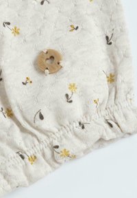 Tissu à motif floral crème avec de petites fleurs jaunes et brunes. Comprend un ourlet froncé et un bouton en bois avec une découpe en forme de cœur.