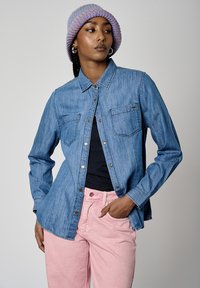 Camisa de denim en azul claro con dos bolsillos en el pecho, botones a presión y costuras en contraste; llevada sobre una blusa negra y combinada con pantalones rosas.