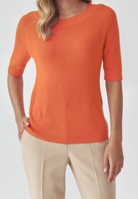 Una donna indossa un maglione a coste arancione a maniche corte con pantaloni beige, una mano in tasca, in piedi davanti a uno sfondo semplice.