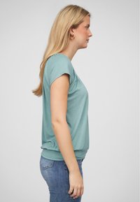 Licht turquoise T-shirt met korte mouwen en een ronde halslijn, met gathered detail bij de taille, gecombineerd met blauwe jeans, weergegeven van opzij.