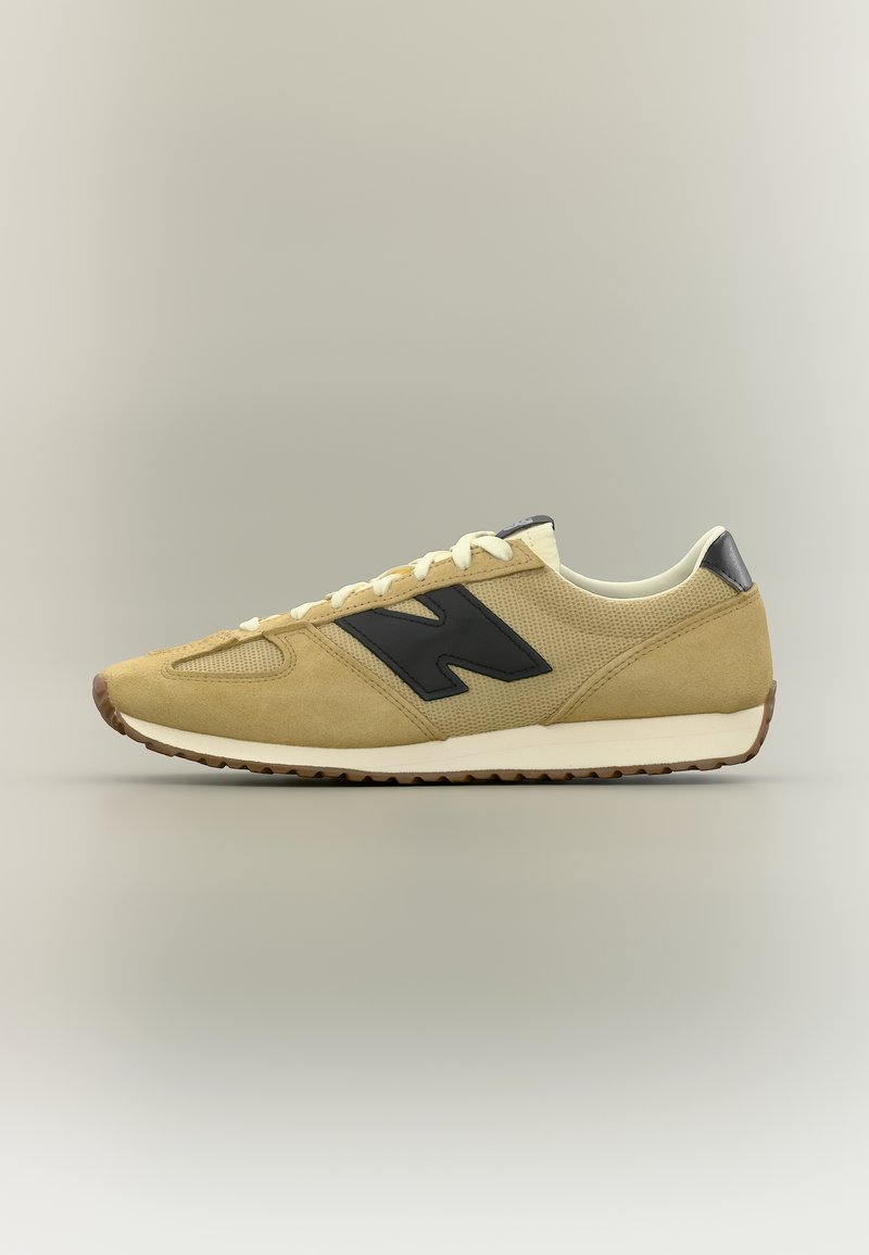 Μπεζ και μαύρο αθλητικό παπούτσι New Balance με μαύρο λογότυπο, λευκά κορδόνια και λευκή σόλα σε ουδέτερο φόντο, εμφανιζόμενο από πλαϊνή όψη.