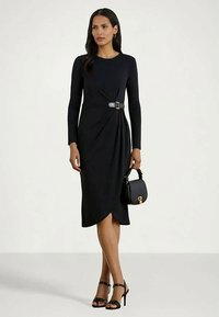 Γυναίκα που φοράει ένα μαύρο midi φόρεμα με μακριά μανίκια και λουρί στο πλάι, κρατώντας μια μαύρη τσάντα και φορώντας μαύρα ψηλοτάκουνα σανδάλια.